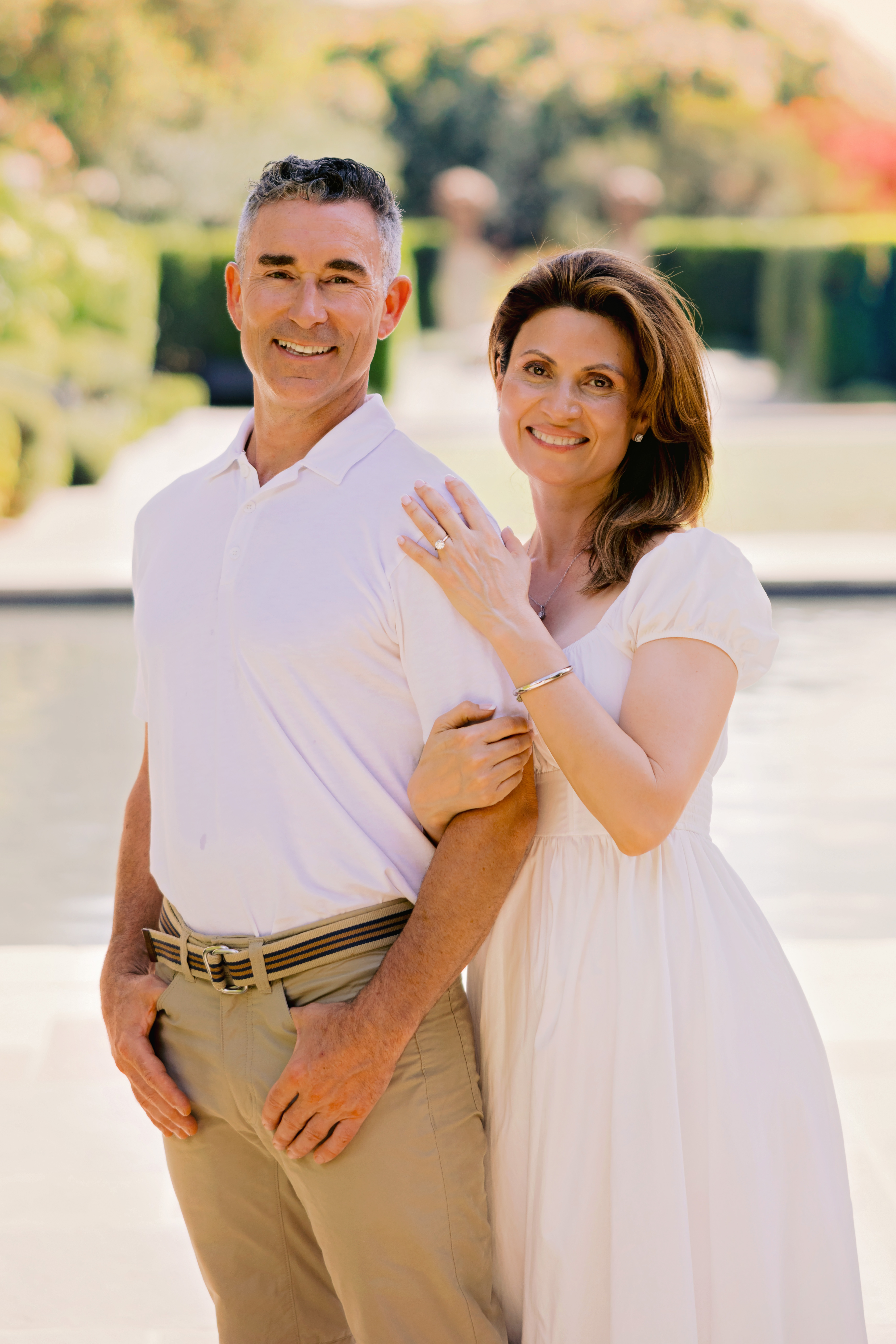 A happy couple poses together outdoors, smiling and showcasing their engagement. The man is wearing a white polo shirt and beige pants, while the woman is dressed in a light-colored dress, displaying her engagement ring. The background features a serene garden setting.